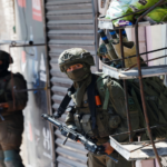 Armed soldiers in tactical gear on a city street, one peering from behind a metal cart stacked with bags adjacent to a shuttered storefront.