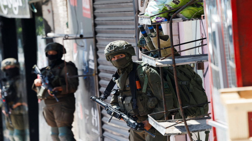 Armed soldiers in tactical gear on a city street, one peering from behind a metal cart stacked with bags adjacent to a shuttered storefront.
