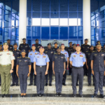 Group of uniformed military officers posing for a formal photo on building steps outside a glass-fronted entrance.
