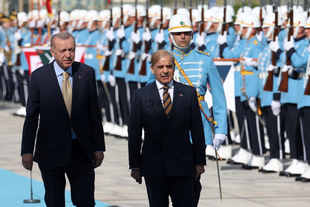 Two men in dark suits walk in front of a ceremonial military parade, with soldiers in blue dress uniforms standing at attention in the background.