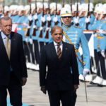 Two men in dark suits walk in front of a ceremonial military parade, with soldiers in blue dress uniforms standing at attention in the background.