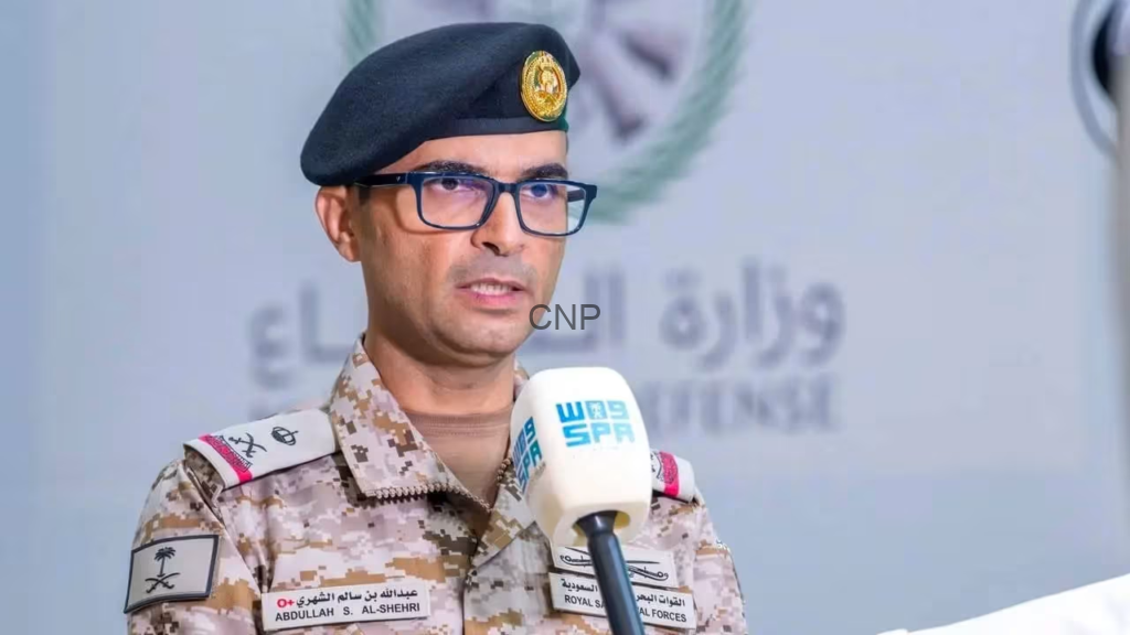 Military officer in desert camouflage uniform and black beret speaking into a handheld mic at a press briefing, wearing glasses.