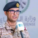 Military officer in desert camouflage uniform and black beret speaking into a handheld mic at a press briefing, wearing glasses.
