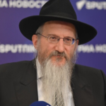Bearded man in a black hat and suit, wearing glasses, speaking at a press conference against a purple backdrop with logos.