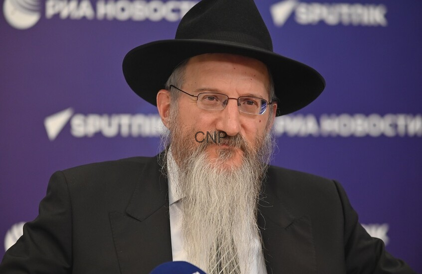 image - Center News Network Bearded man in a black hat and suit, wearing glasses, speaking at a press conference against a purple backdrop with logos.