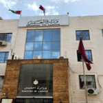 Front view of a government building labeled Ministry of Labour, with large glass windows, palm trees, and flagpoles outside; Arabic and English signage visible.