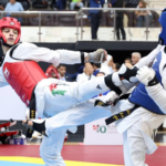 Two taekwondo fighters in red and blue protective gear exchange kicks on a competition mat as spectators watch behind them.