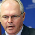 Close-up of a man with gray hair and glasses in a dark suit, speaking at a press event against a blue backdrop.