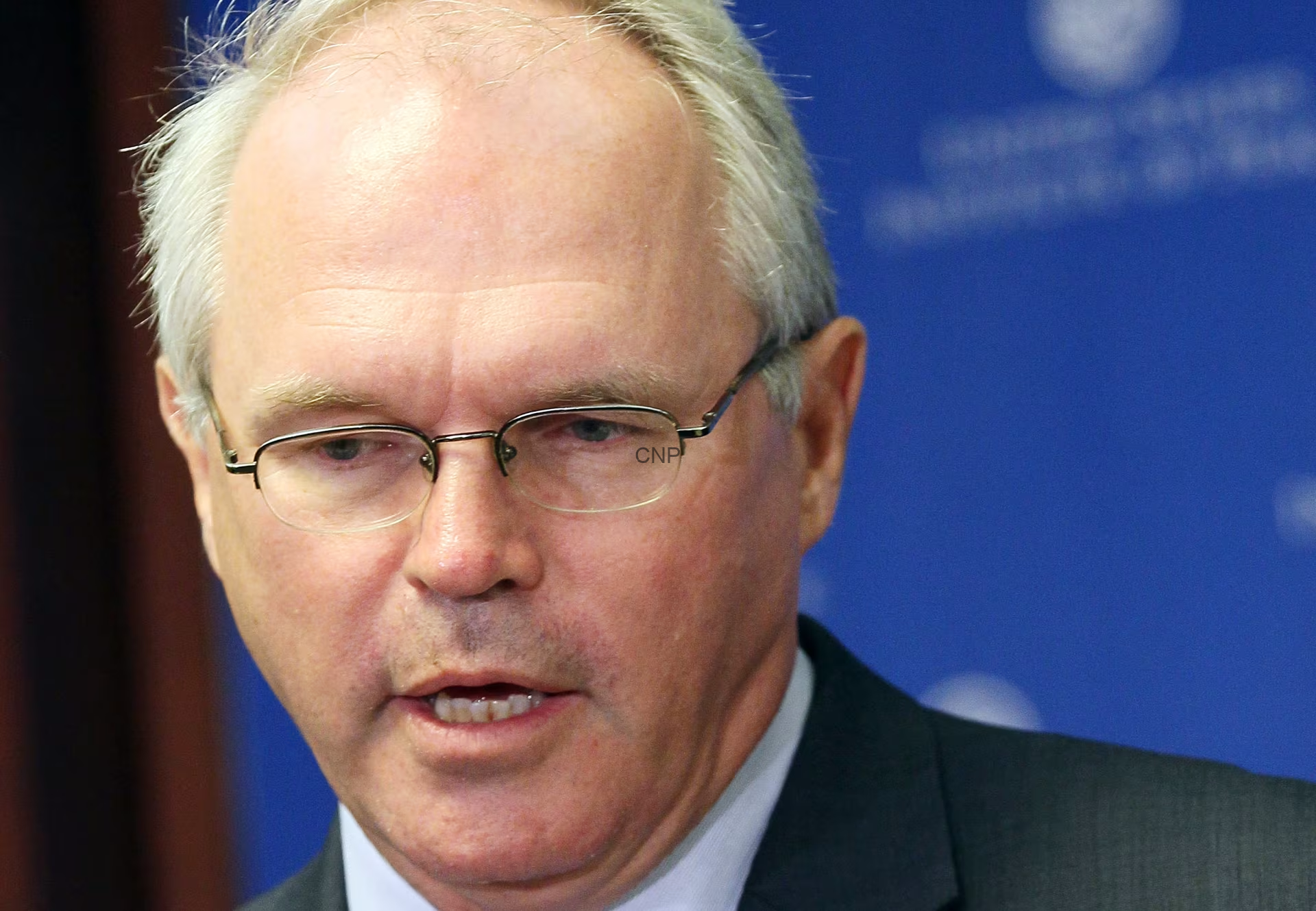 Close-up of a man with gray hair and glasses in a dark suit, speaking at a press event against a blue backdrop.