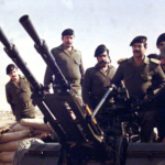 Soldiers in uniform man large mounted artillery guns on a military vehicle in a desert with sandbags nearby.