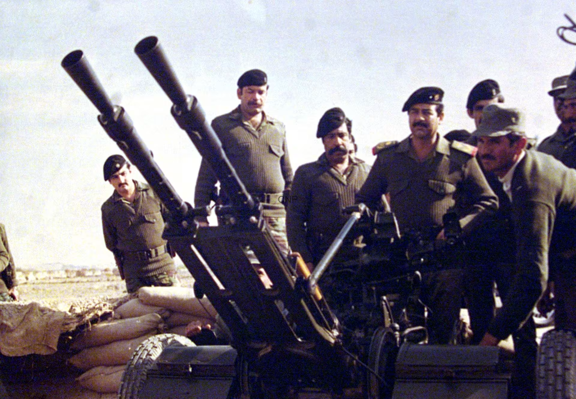 Soldiers in uniform man large mounted artillery guns on a military vehicle in a desert with sandbags nearby.