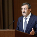 Man in a dark suit delivering a speech at a wooden podium with two microphones, Turkish flag in the background.