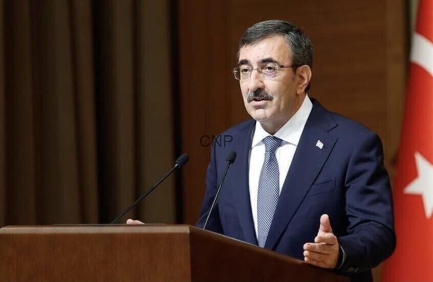 image - Center News Network Man in a dark suit delivering a speech at a wooden podium with two microphones, Turkish flag in the background.