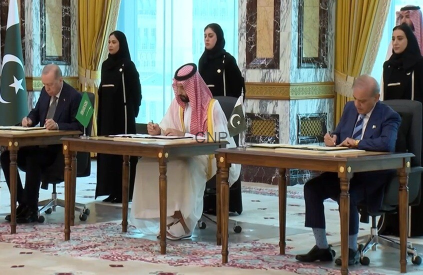 image - Center News Network Bilateral signing ceremony between Pakistan and Saudi Arabia with officials signing documents at desks, flanked by flag-bearing aides in an ornate room.