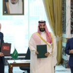 Three men in formal attire hold folders during a signing ceremony; Turkish and Saudi flags on the table, ornate room backdrop.