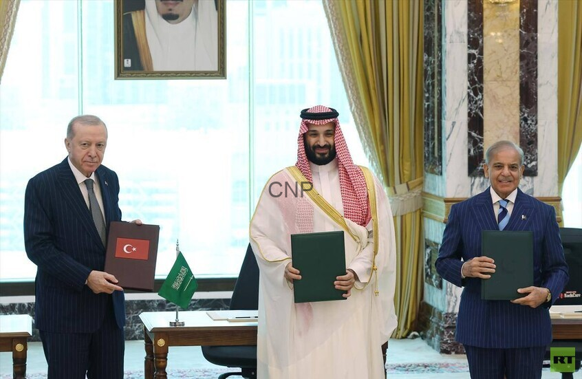 image - Center News Network Three men in formal attire hold folders during a signing ceremony; Turkish and Saudi flags on the table, ornate room backdrop.