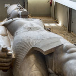 Massive reclining statue of an ancient Egyptian pharaoh inside a museum lobby, seen from above with one arm extended.