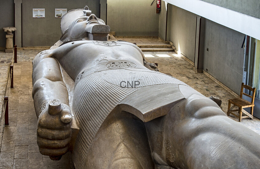 Massive reclining statue of an ancient Egyptian pharaoh inside a museum lobby, seen from above with one arm extended.
