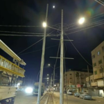 Night street scene with tall utility poles and overhead power lines, glowing streetlights, and parked cars along the road.