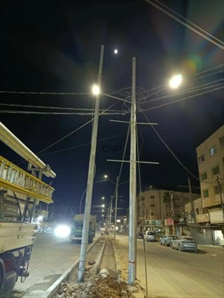 Night street scene with tall utility poles and overhead power lines, glowing streetlights, and parked cars along the road.