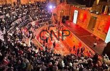 image - Center News Network Large crowd gathered around a nighttime outdoor stage with red lights, watching a performance.