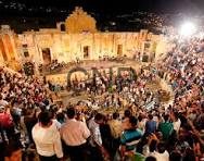image - Center News Network Large night-time crowd gathered in a historic plaza, standing around a circular ground design, with a bright, ornate building illuminated in the background.