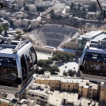 Two cable cars swing along a cable above an urban landscape, with a large circular stadium visible below.