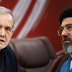 Two men sit together in a serious discussion, one with gray hair in a dark shirt and the other with a black turban and glasses wearing traditional attire in front of a red backdrop.