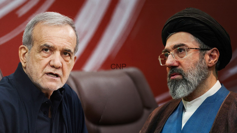 Two men sit together in a serious discussion, one with gray hair in a dark shirt and the other with a black turban and glasses wearing traditional attire in front of a red backdrop.