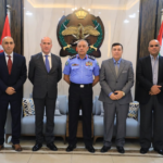 Five men in formal attire pose for a group photo in a government lobby, center officer in blue uniform with medals, emblem and flags in the background.