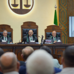 A panel of three judges in robes sits at a raised bench in a courtroom, with a flag on the left and a large emblem behind them.