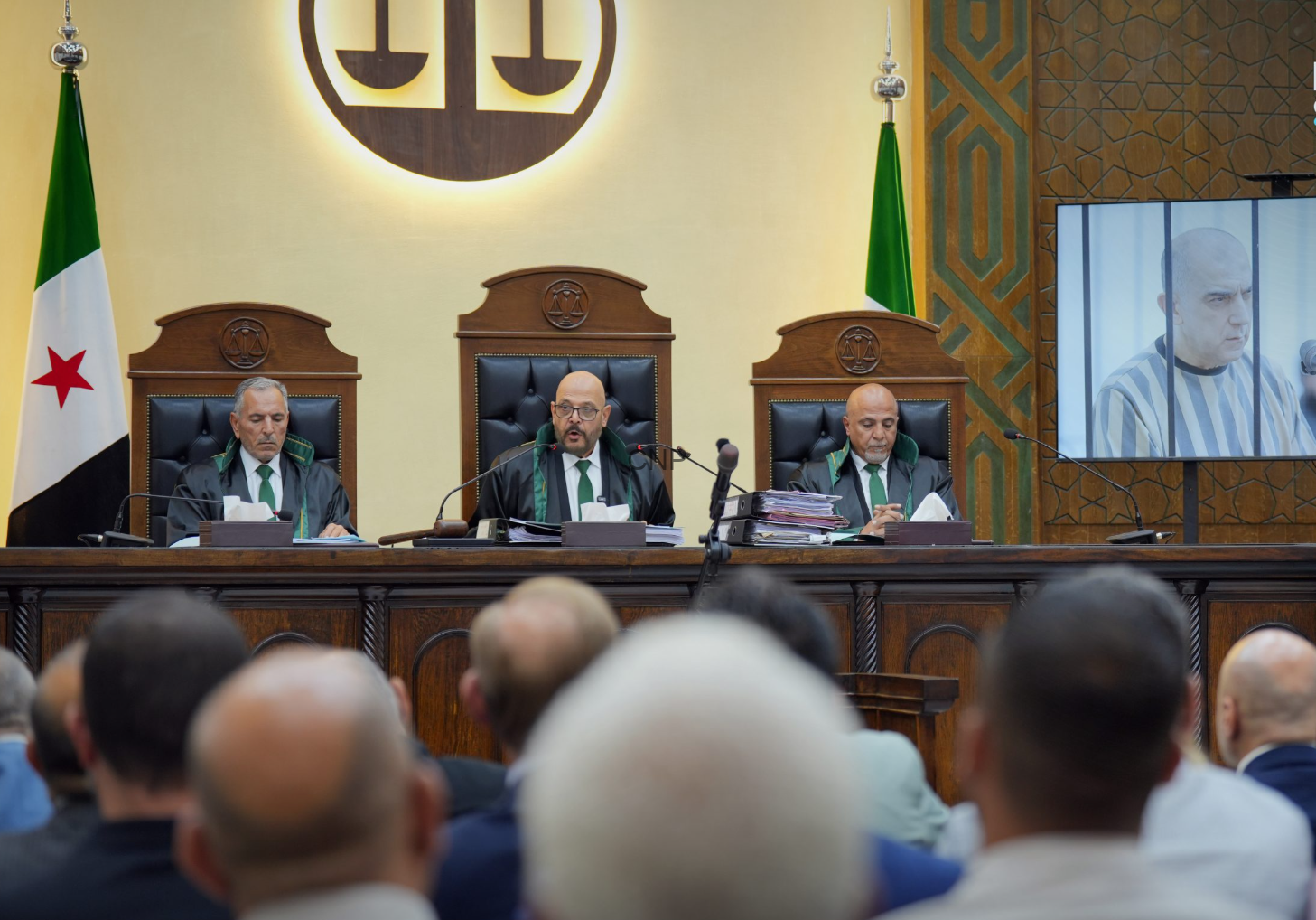 A panel of three judges in robes sits at a raised bench in a courtroom, with a flag on the left and a large emblem behind them.