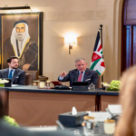 Two men in suits at a formal meeting table, one gesturing while a flag stands nearby, with a large portrait on the wall behind.