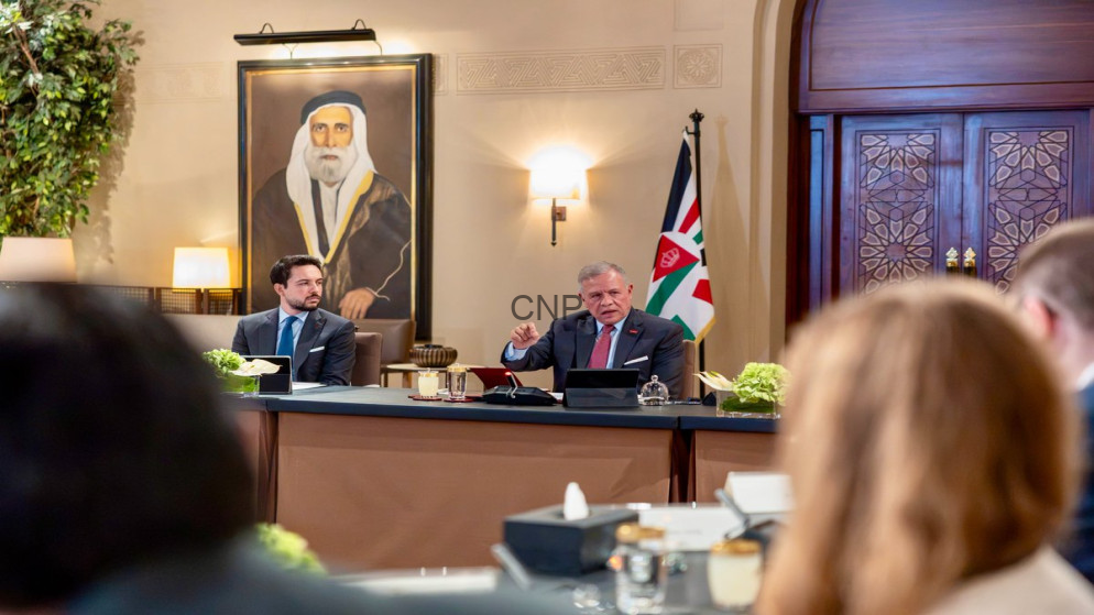 image - Center News Network Two men in suits at a formal meeting table, one gesturing while a flag stands nearby, with a large portrait on the wall behind.