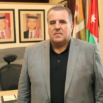 Man in a gray suit stands in an office with framed portraits on the wall and flags in the background.