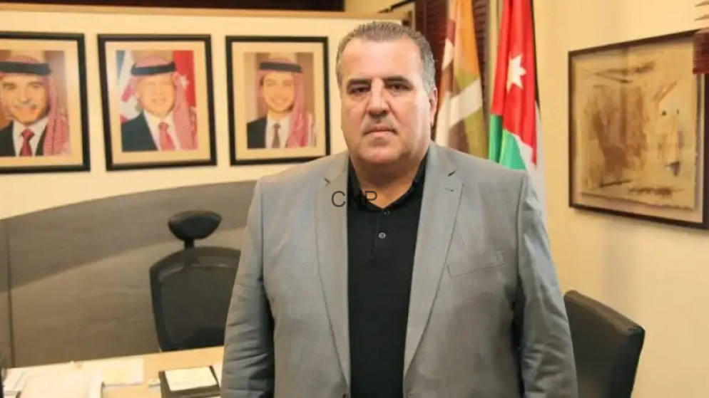 Man in a gray suit stands in an office with framed portraits on the wall and flags in the background.