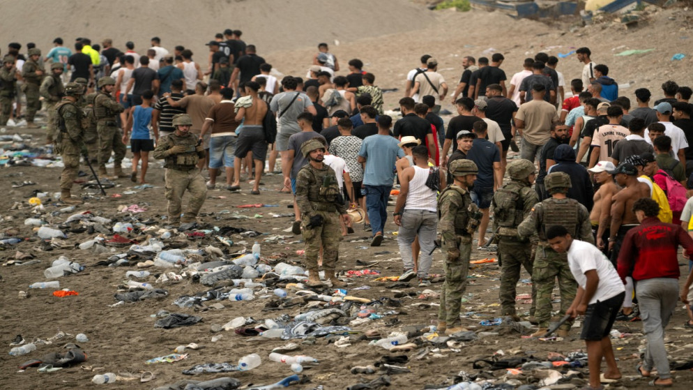 image - Center News Network Armed soldiers stand on a litter-strewn beach as a crowd of civilians forms a long line behind them.