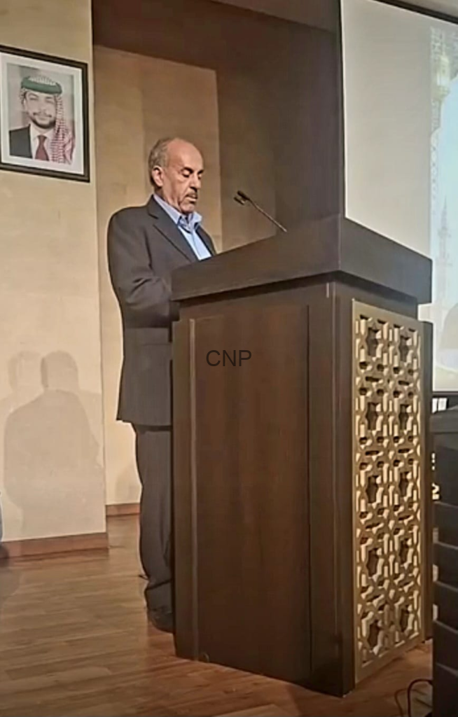 An older man in a suit standing at a wooden podium, speaking into a microphone in a conference room.