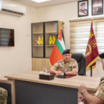 Three military officers in camouflage sit around a large desk in a formal office, with flags and framed portraits in the background.