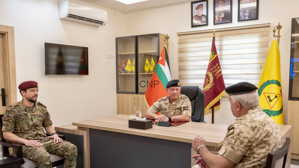 Three military officers in camouflage sit around a large desk in a formal office, with flags and framed portraits in the background.