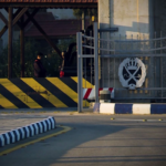 Gated security entrance with a decorative crest on the gate and a black-and-yellow striped barrier along a curved road, with a person in the background.