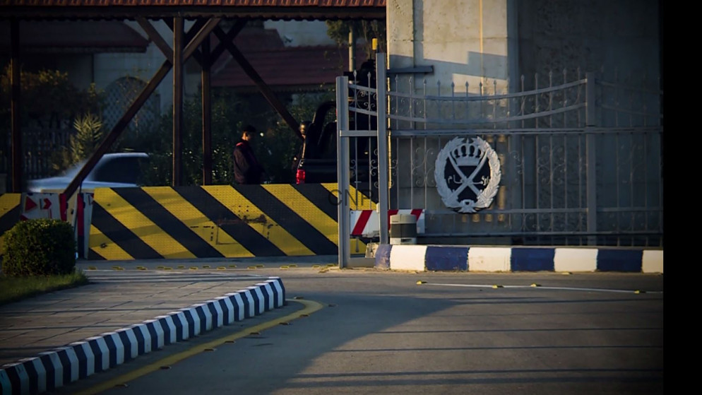 image - Center News Network Gated security entrance with a decorative crest on the gate and a black-and-yellow striped barrier along a curved road, with a person in the background.