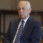 Older man in a dark suit and striped tie seated in a formal setting with a serious expression.