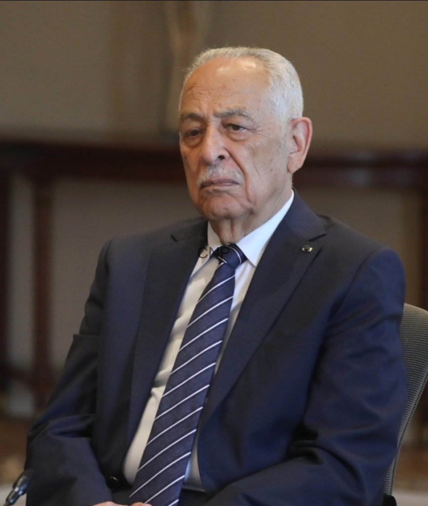 image - Center News Network Older man in a dark suit and striped tie seated in a formal setting with a serious expression.