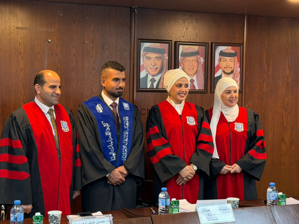 Four graduates in ceremonial gowns (red and blue sashes) stand side by side in a formal room with wood-paneled walls and framed portraits behind them.