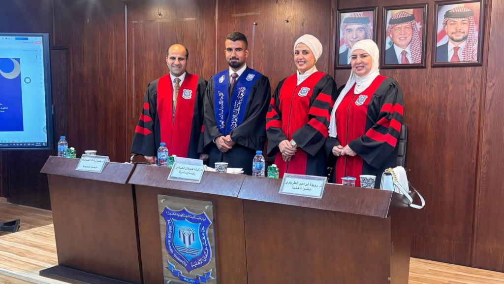 image - Center News Network Four people in red-and-black academic robes stand behind a desk in a university conference room, with framed portraits on a wood-paneled wall behind them.