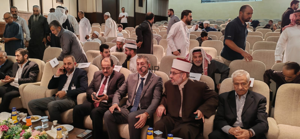 Men in suits and traditional attire seated and standing in a large auditorium, conversing near a front-row table with drinks and snacks.