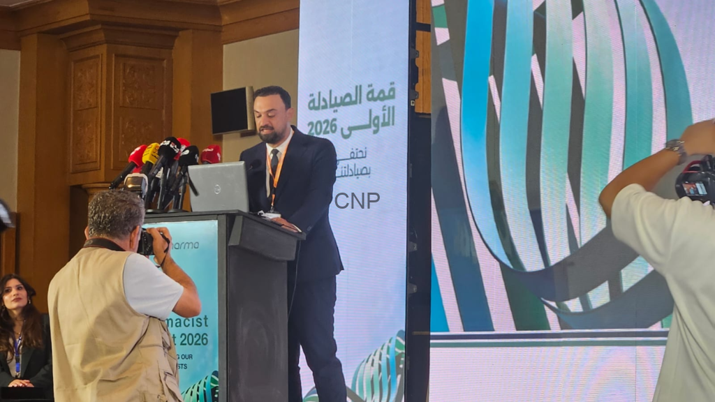 Man in a suit giving a speech at a podium with multiple microphones, a large screen with Arabic text behind him, and photographers in the foreground.