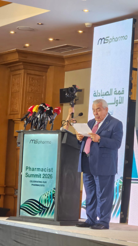 Man in a navy suit reading from a sheet at a podium with multiple microphones during Pharmacist Summit 2026; branded display behind him.