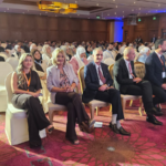 Group of professionals in business attire seated in rows at a conference hall, wearing lanyards and facing the stage.
